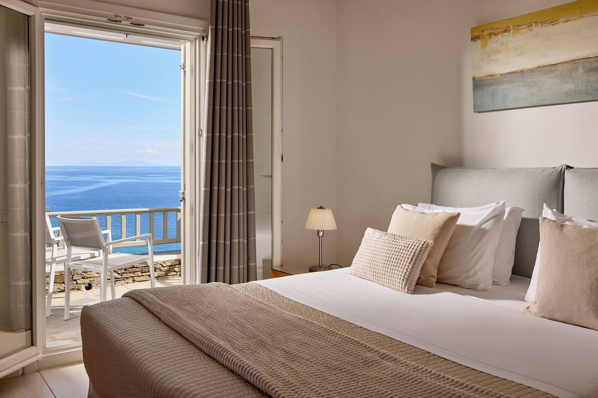 A hotel room with a bed, a nightstand, a lamp, and a view of the sea.