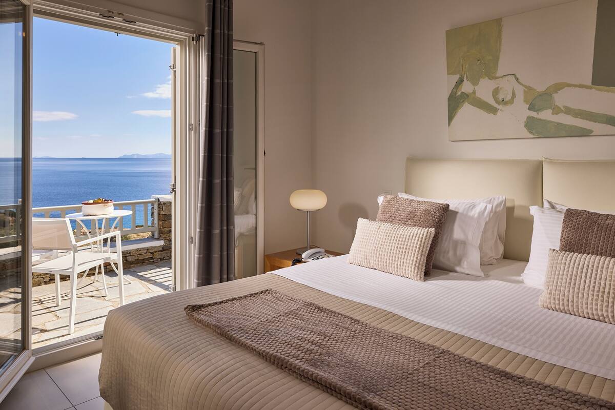 A bedroom with a large bed, a nightstand, a lamp, and a view of the sea through a sliding glass door.