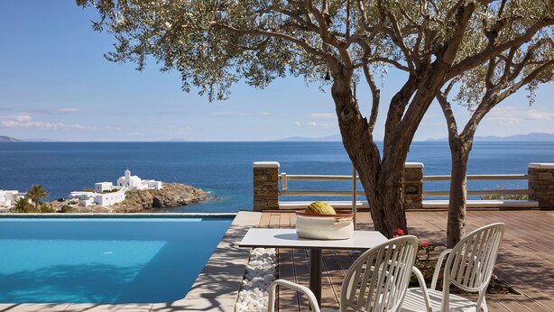A poolside area with a table and chairs, overlooking a clear blue sea and white buildings in the distance. - Selana Suites (Sifnos)