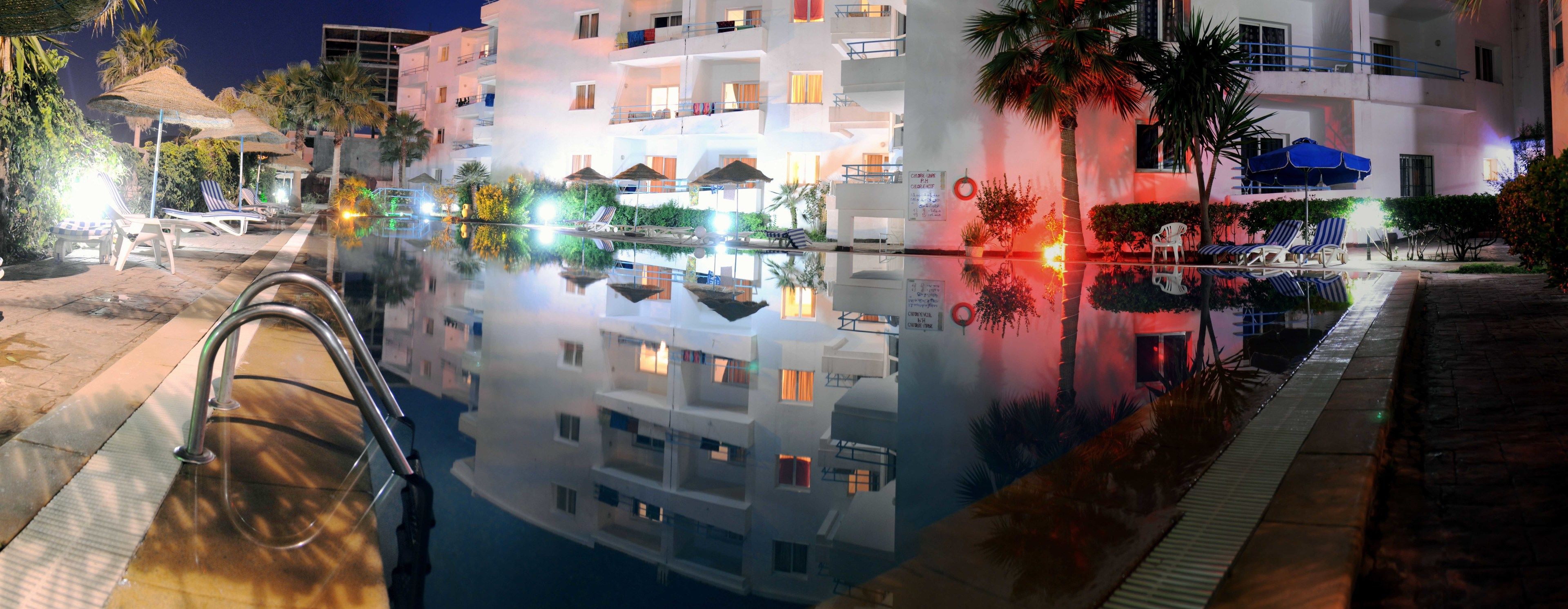 A hotel with a pool reflecting the building and palm trees.