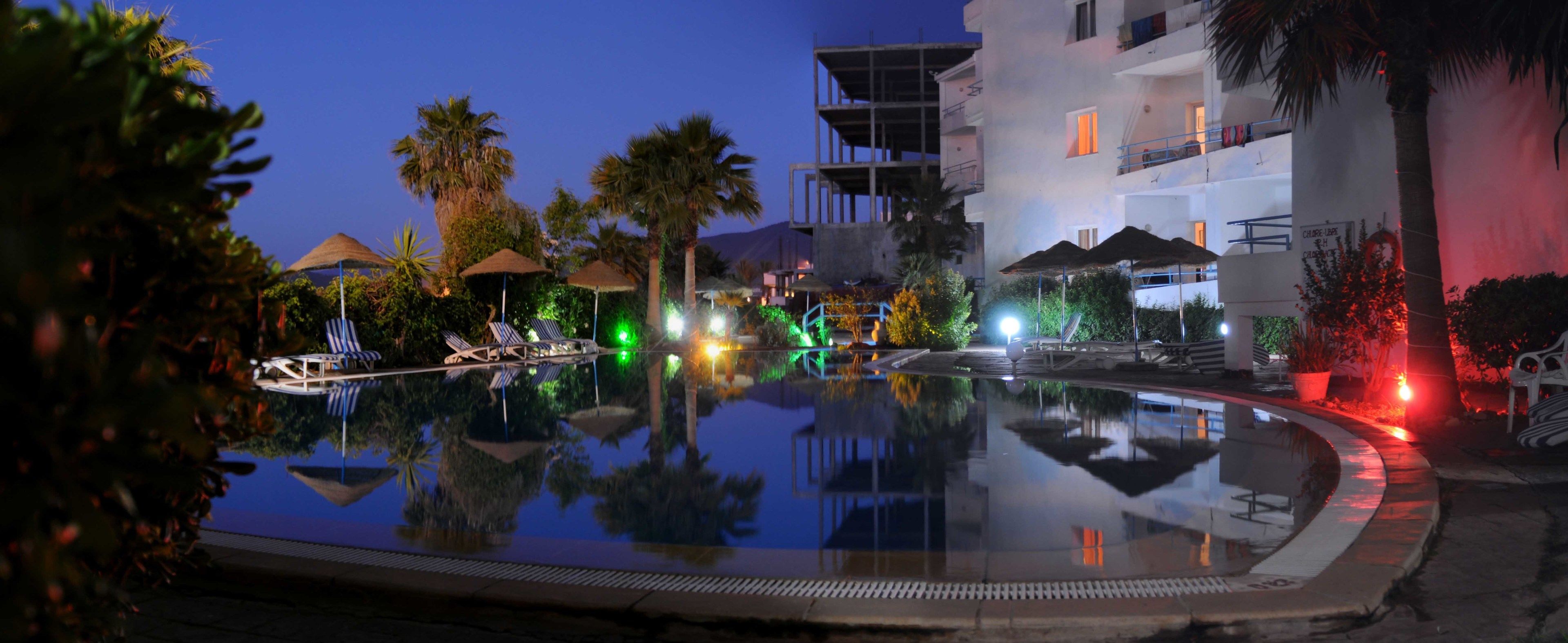 Vista nocturna d'una zona de piscina d'hotel amb gandules i ombrel·les, envoltada de palmeres i un edifici amb balcons.