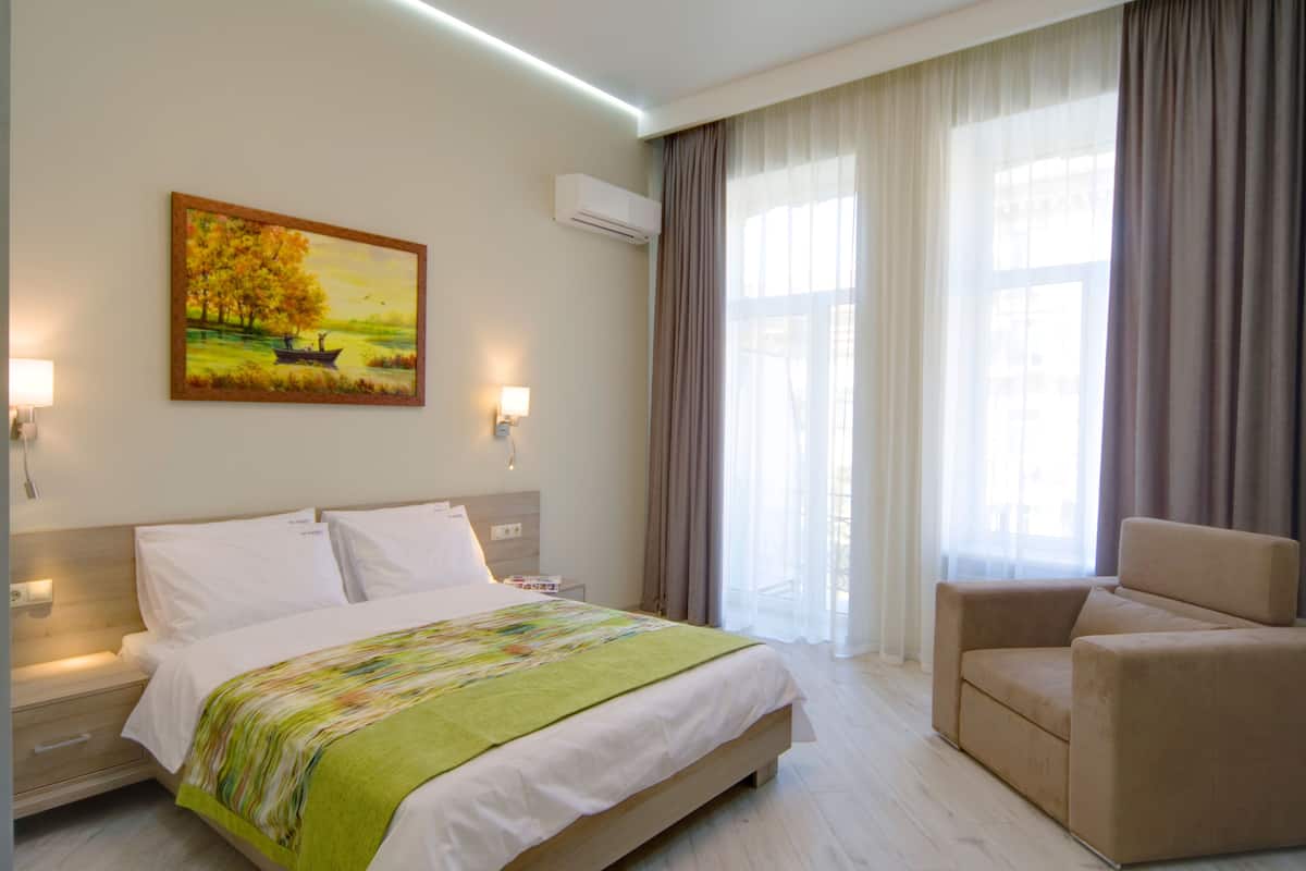 A hotel room with a bed, a chair, a painting on the wall, and a window with curtains.