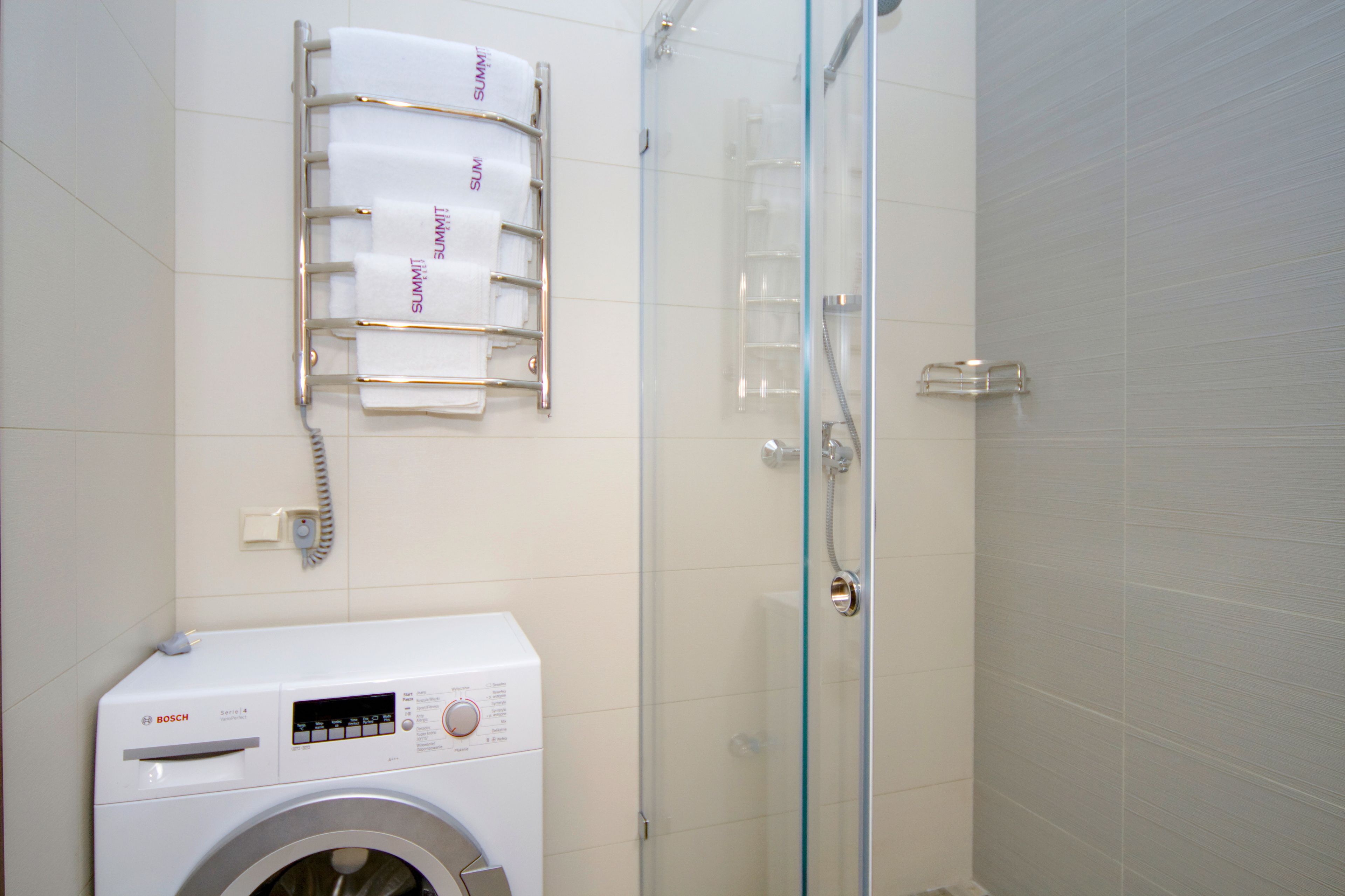 A modern bathroom with a Bosch washing machine, a glass shower enclosure, and a towel rack with towels labeled "SUMMIT".