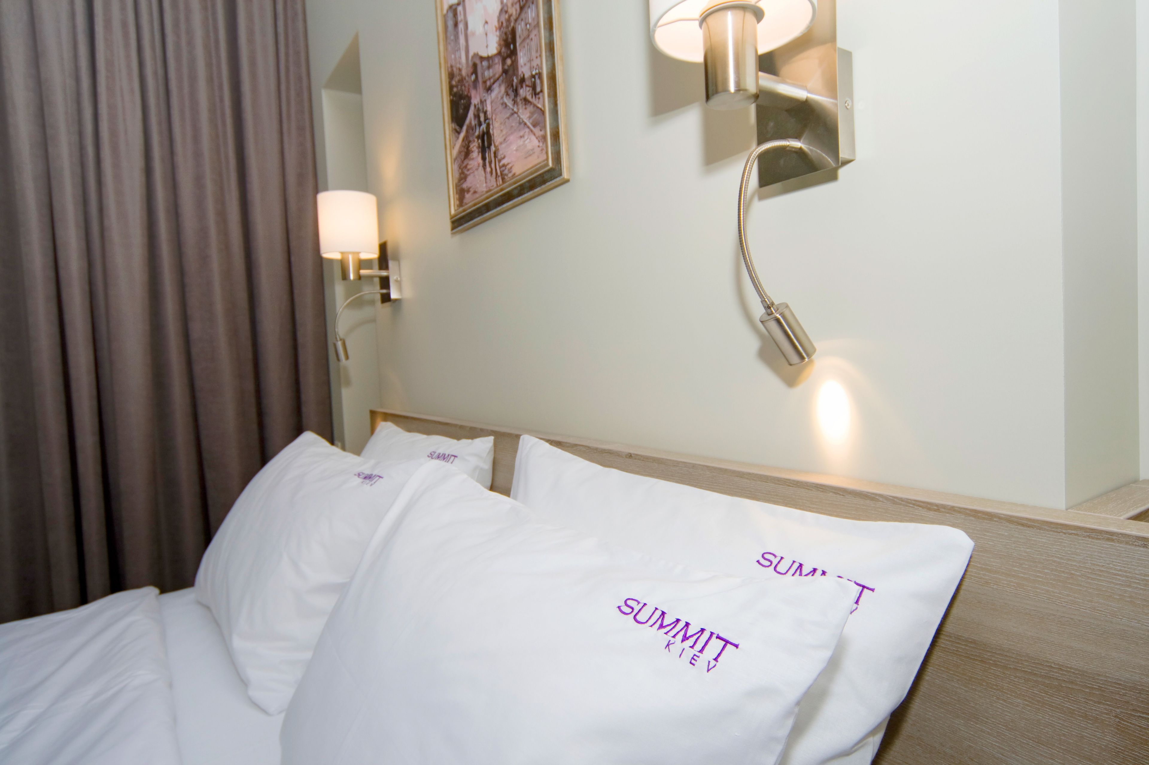 A hotel room with a bed, white pillows with "SUMMIT" text, a wall-mounted lamp, and a painting on the wall.