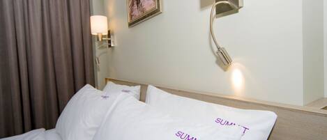 Quarto de hotel com cama, almofadas brancas com o texto "SUMMIT" e uma luminária fixada na parede, além de um quadro na parede.