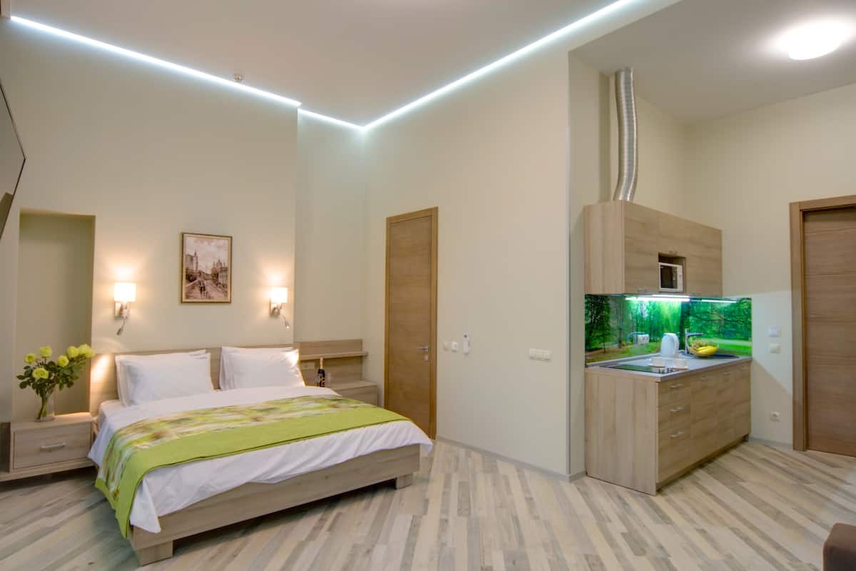 A bedroom with a bed, bedside tables, a nightstand, a wall-mounted lamp, a picture, and a built-in kitchen area with a microwave and sink.