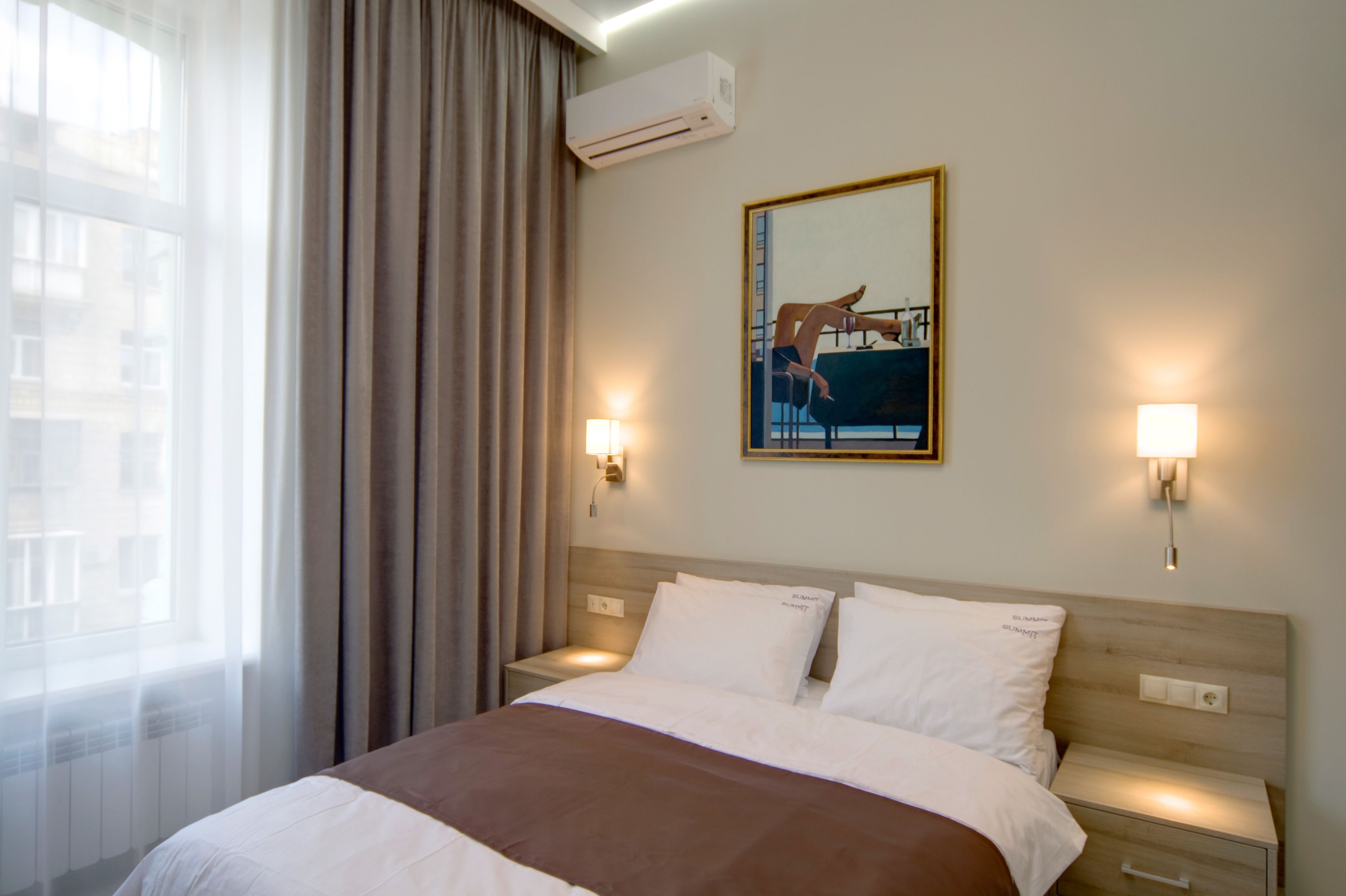 Une chambre d’hôtel comprenant un lit, deux tables de chevet, une commode, un climatiseur fixé au mur, une fenêtre avec des rideaux et deux lampes fixées au mur.
