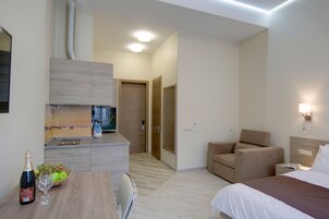 Una habitación de hotel moderna con cama, zona de estar, pequeña kitchenette y mesa de comedor con una botella de champán y fruta.
