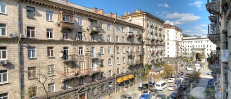A multi-story building with balconies, a street with parked cars, and a clear blue sky.