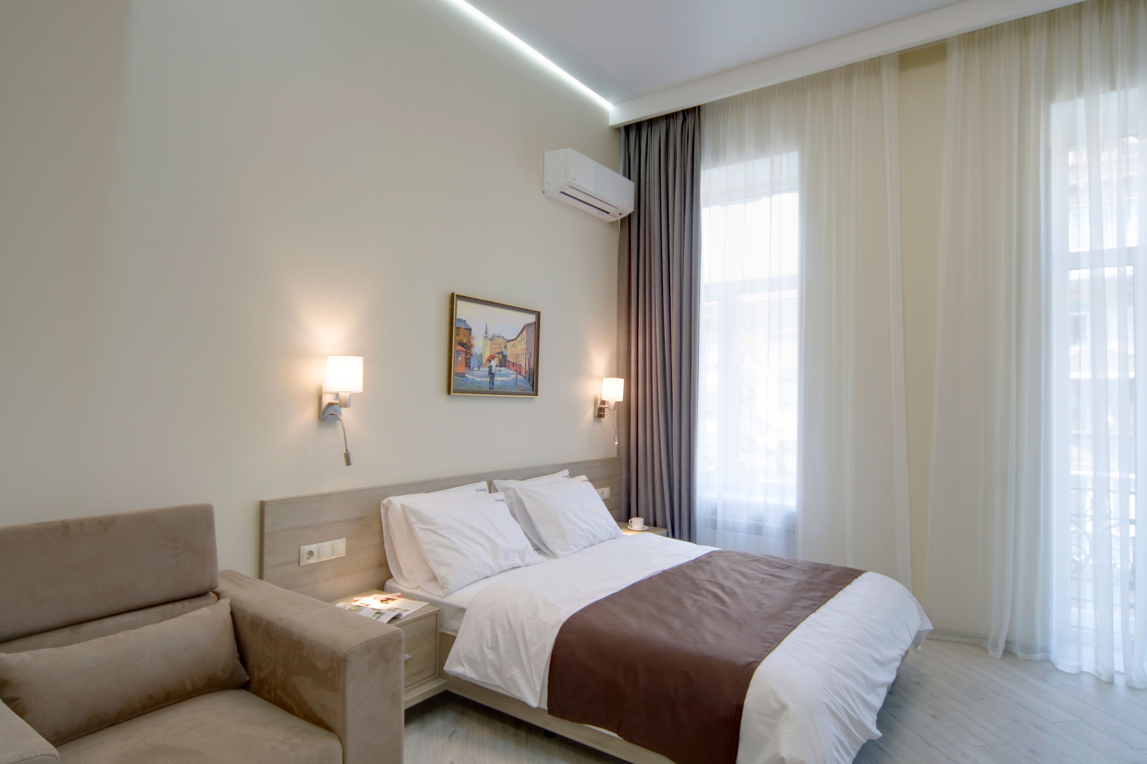 A hotel room with a bed, a chair, a painting on the wall, and an air conditioning unit.