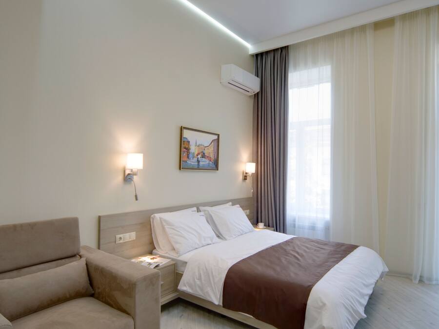 A hotel room with a bed, a chair, a painting on the wall, and an air conditioning unit.