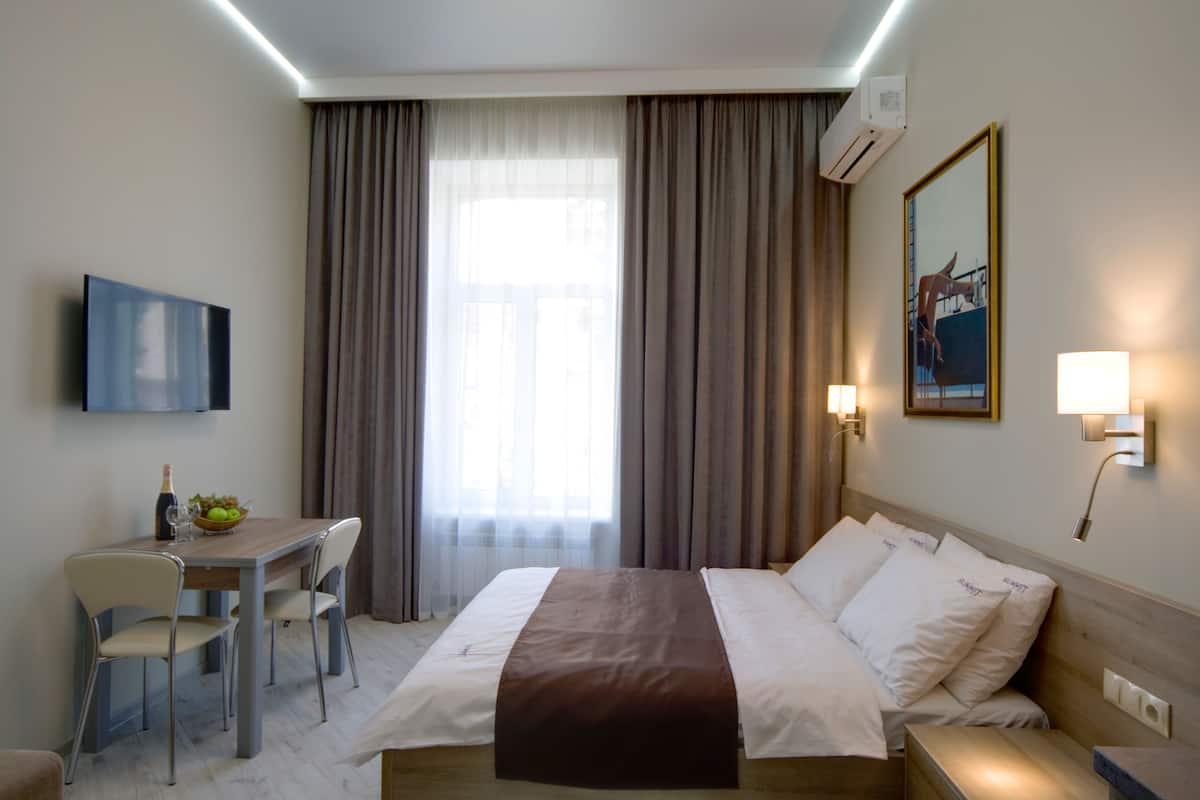 A hotel room with a bed, bedside table, lamp, TV, and a desk with a chair.