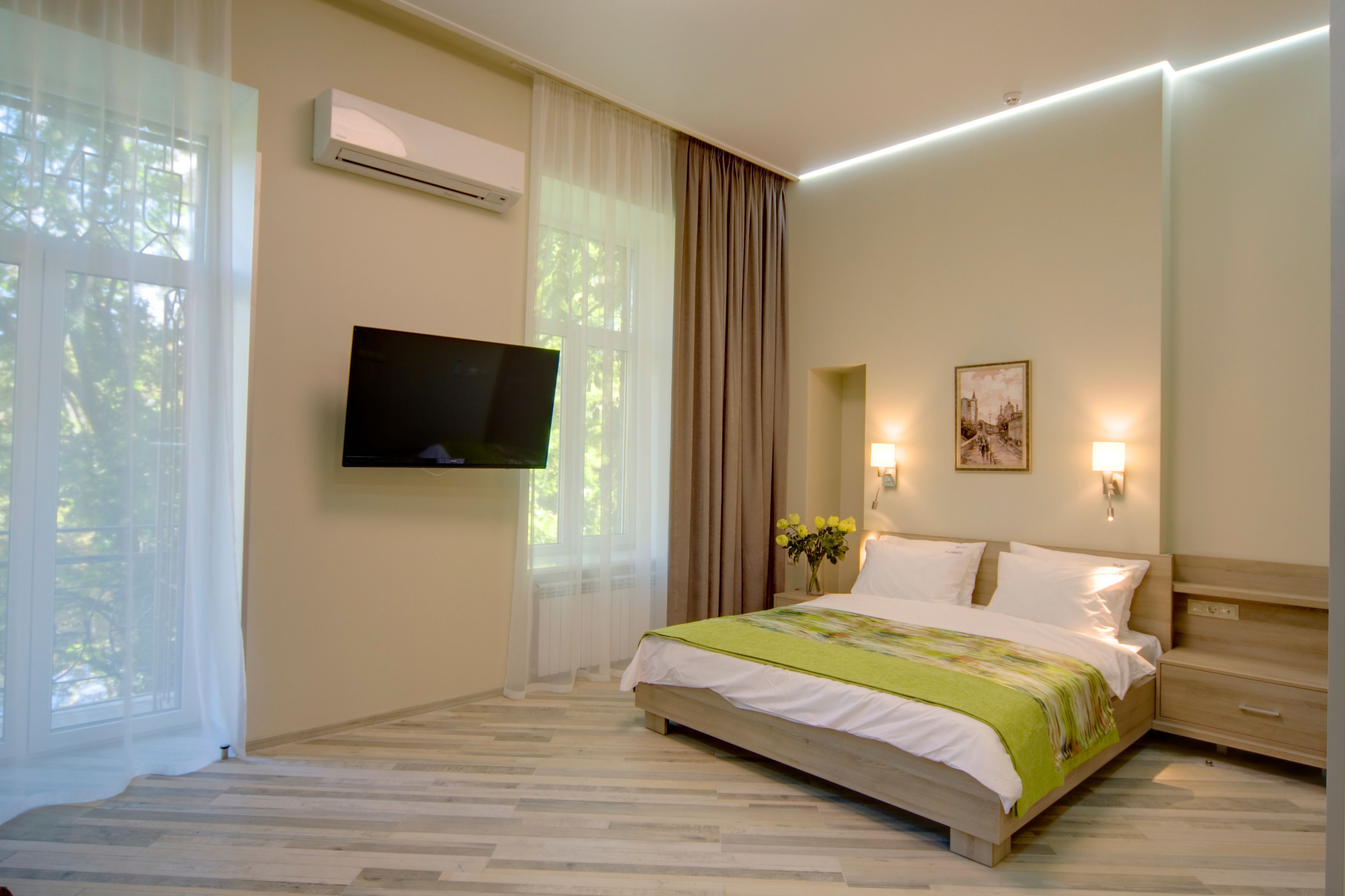 A hotel room with a bed, a flat-screen TV mounted on the wall, a bedside table with a lamp, and a window with sheer curtains.