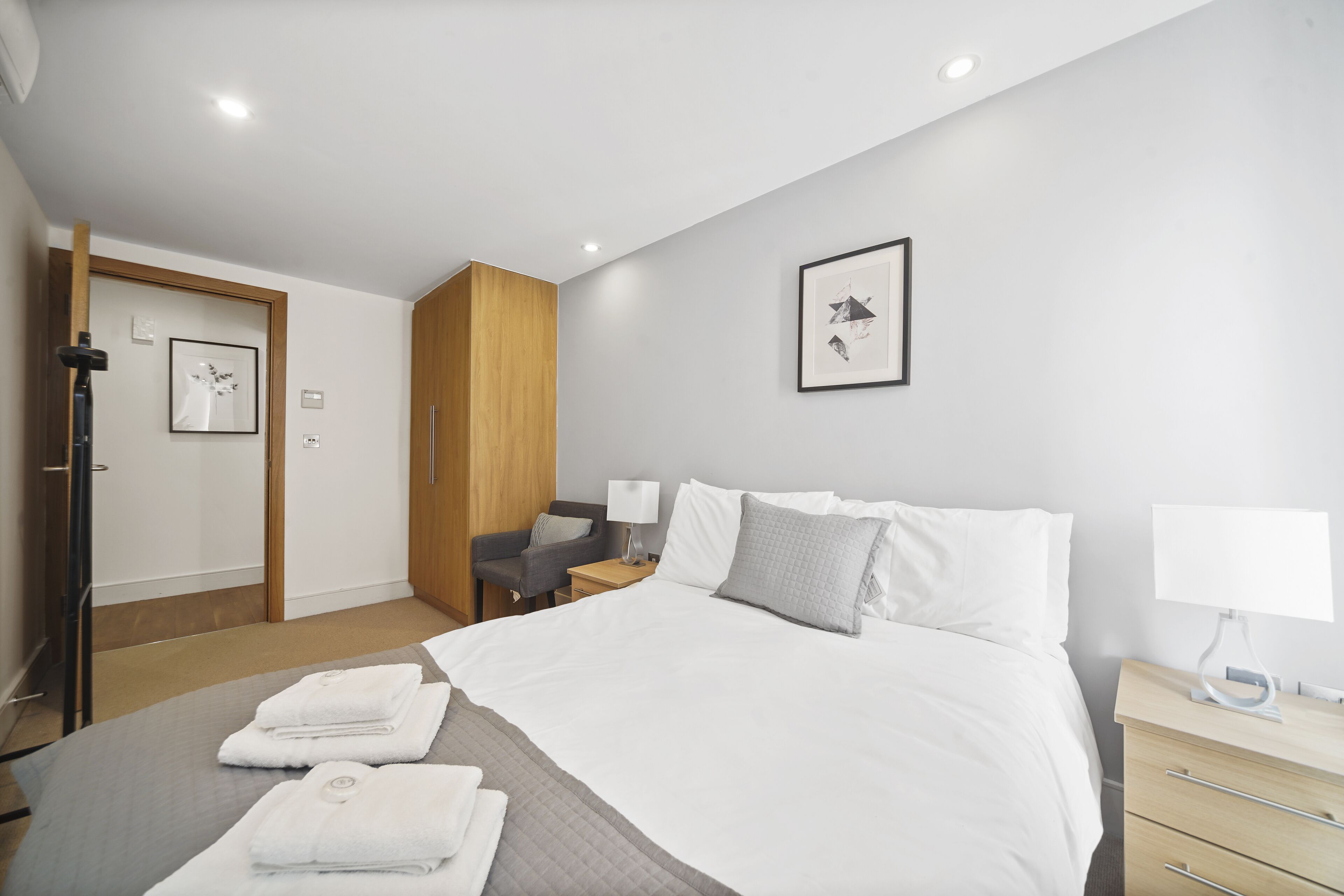 Une chambre d’hôtel moderne avec un lit soigneusement fait, une armoire en bois, un bureau avec une lampe et un tableau encadré au mur.