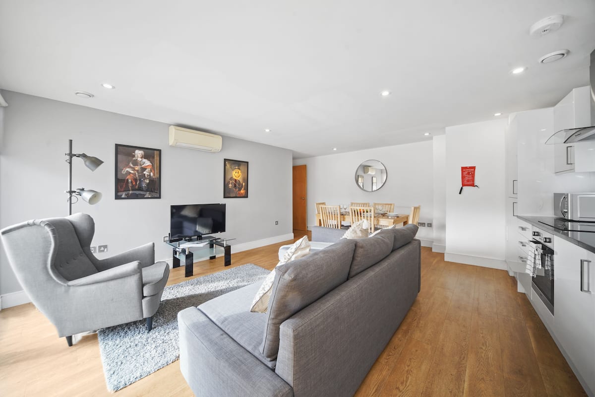 A modern living room with a grey sofa, a grey armchair, a flat-screen TV, and a dining area with wooden chairs and a table.