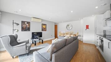 Een moderne woonkamer met een grijze bank, een grijze fauteuil, een flatscreen televisie en een eethoek met houten stoelen en een tafel.