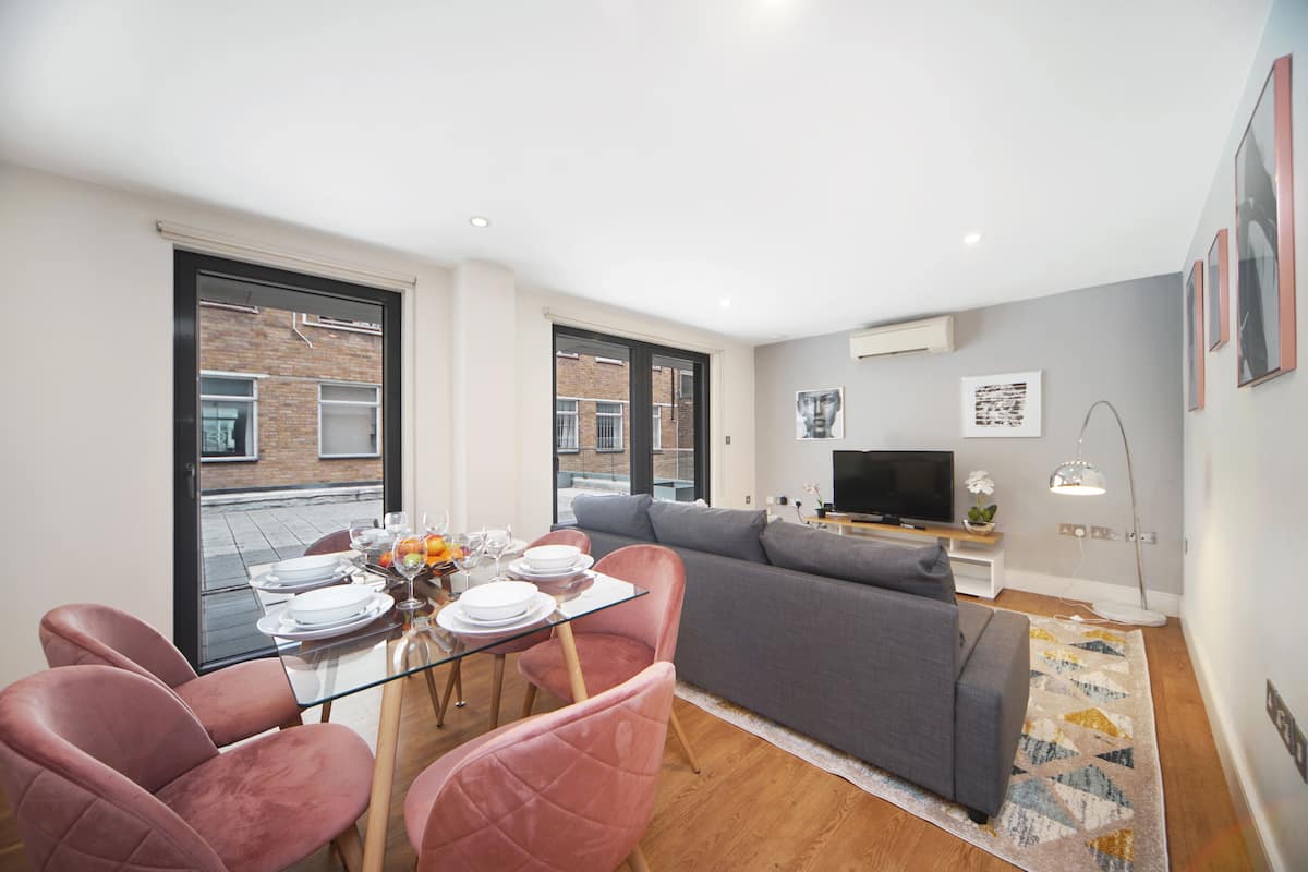 A modern living room with a grey sofa, a glass dining table with pink chairs, and a TV on a stand.