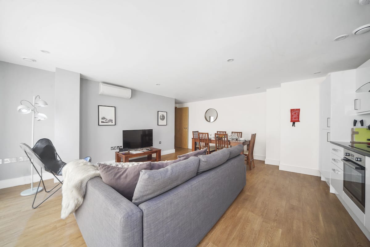 A modern living room with a grey sofa, a dining table with chairs, a flat-screen TV, and a kitchen area with white cabinets and a stove.
