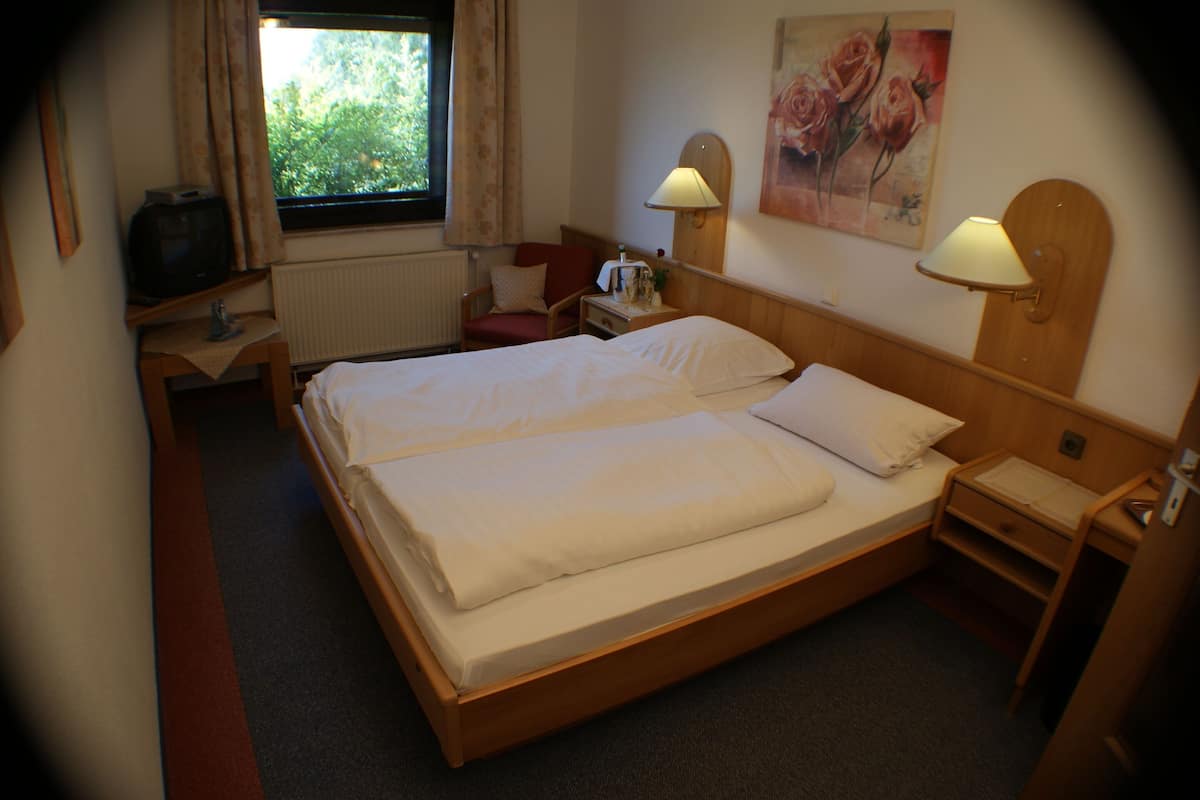 A hotel room with a double bed, a bedside table with a lamp, a chair, a small table with a radio, and a window with curtains.
