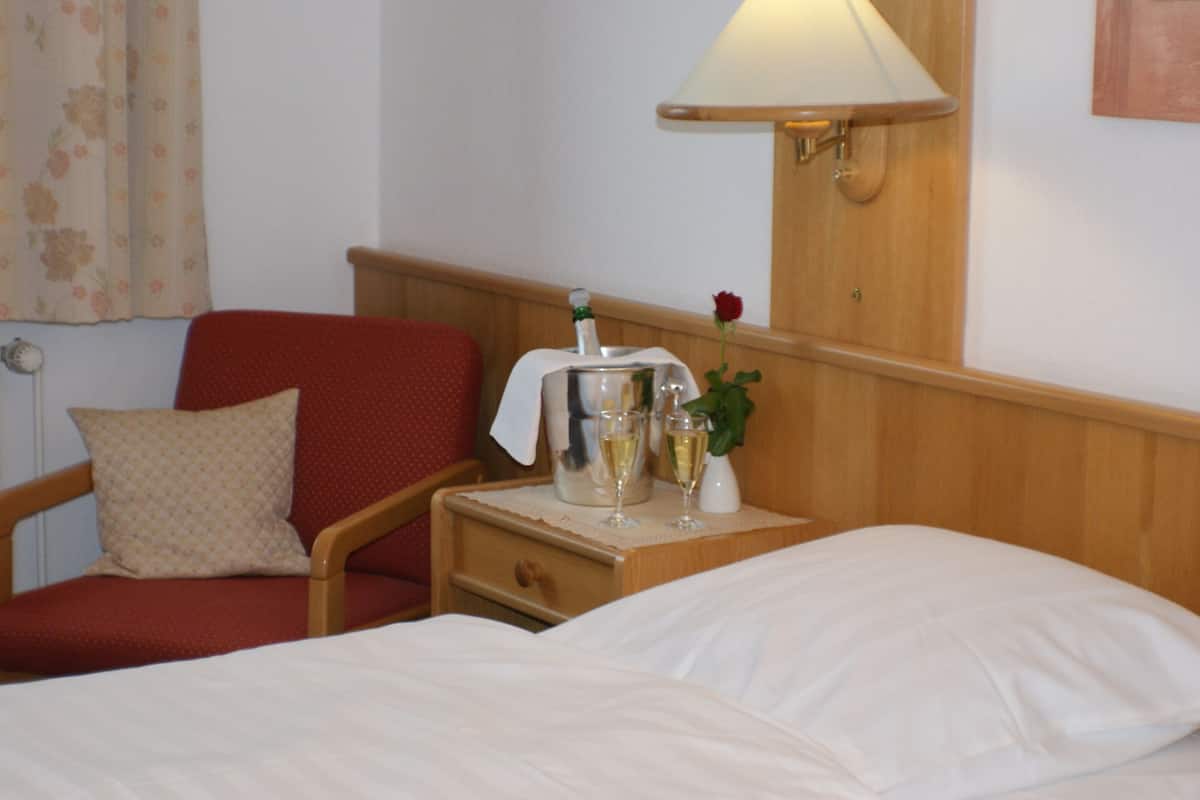 A hotel room with a bed, a wooden nightstand, a red armchair, a small table with a bottle, glasses, and a vase with a rose.