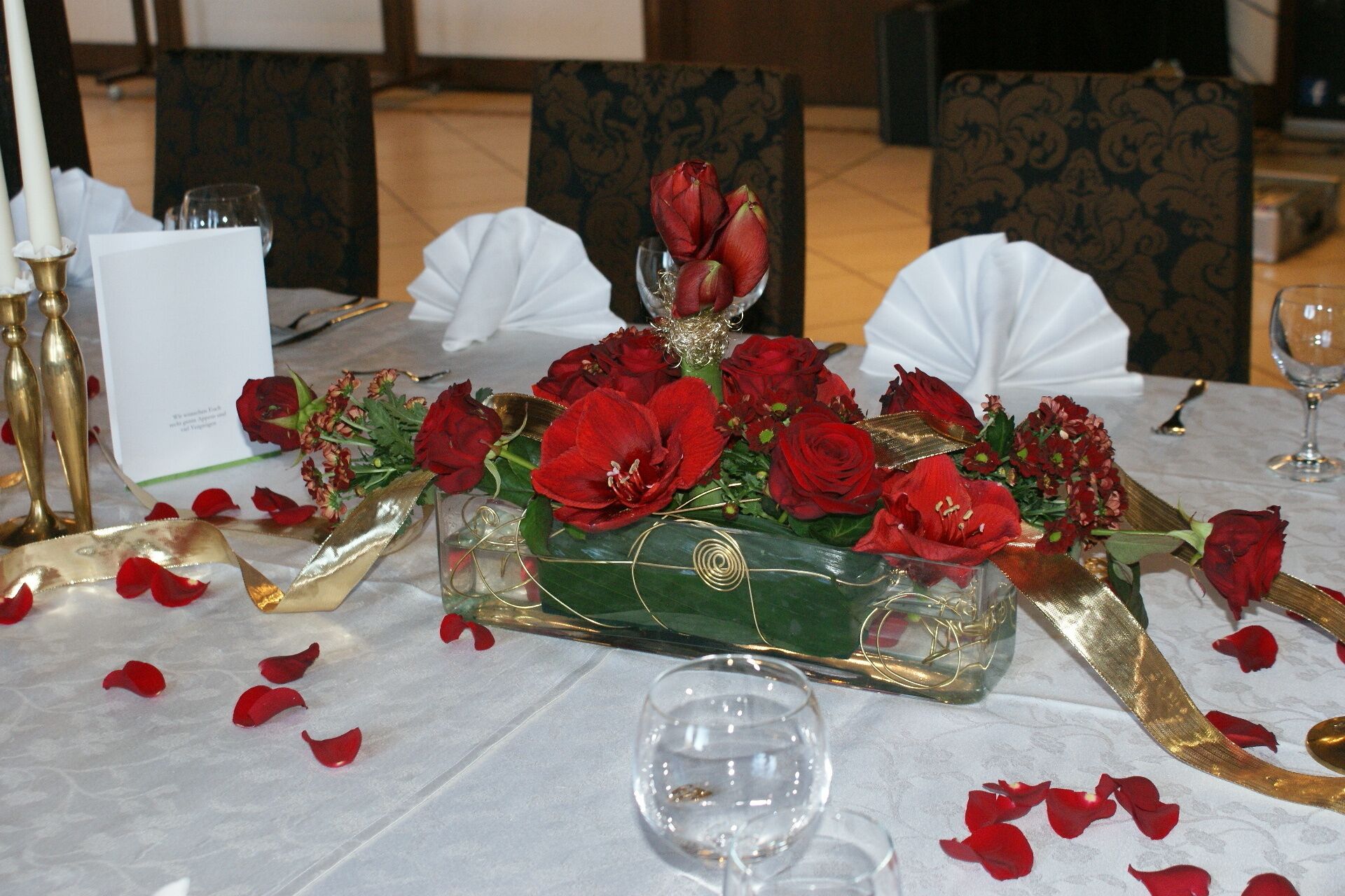 A table set for a formal event with a centerpiece of red roses, a glass vase, and gold accents.