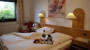 Ein Hotelzimmer mit zwei Betten, einem fensterbelaubten Fenster, einem Rosenbild, zwei Nachttischlampen, einem Tablett mit Frühstücksgebäck und einer Heizung.