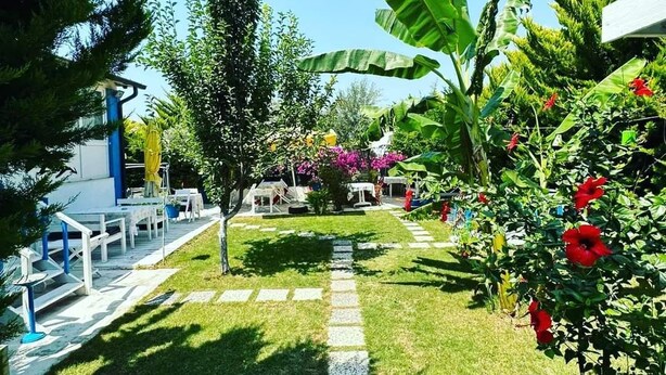 A garden pathway leading to a seating area with tables and chairs, surrounded by lush greenery and flowering plants. - Hotel Antonio (Cesme)
