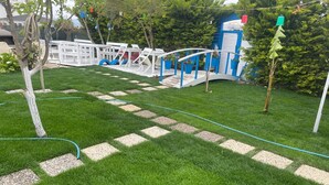 A well-maintained garden area with a paved pathway, outdoor seating, and a small structure with a blue door. - Hotel Antonio (Cesme)