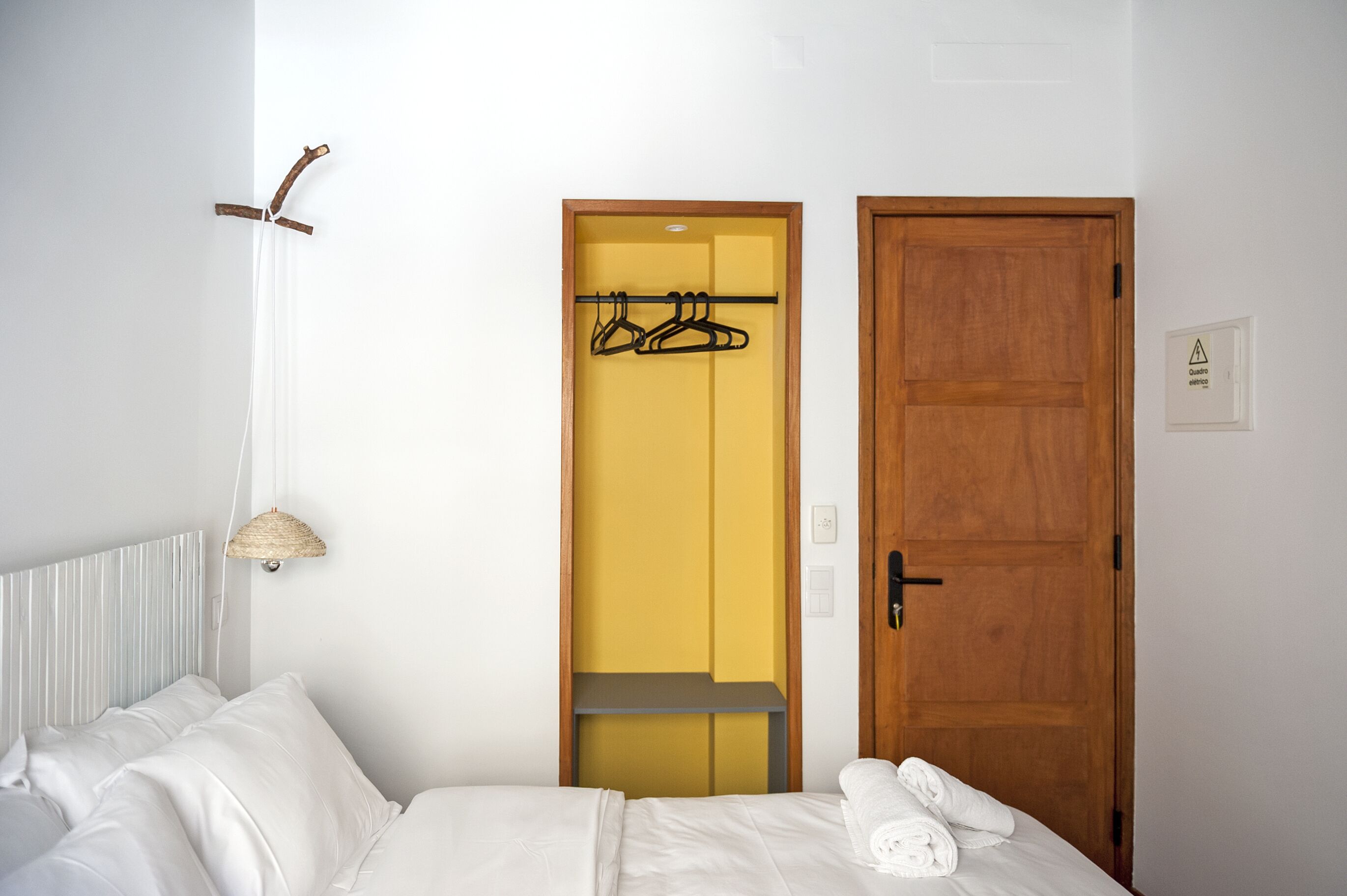 A hotel room with a bed, a hanging light fixture, a wooden door, and a coat rack with hangers.