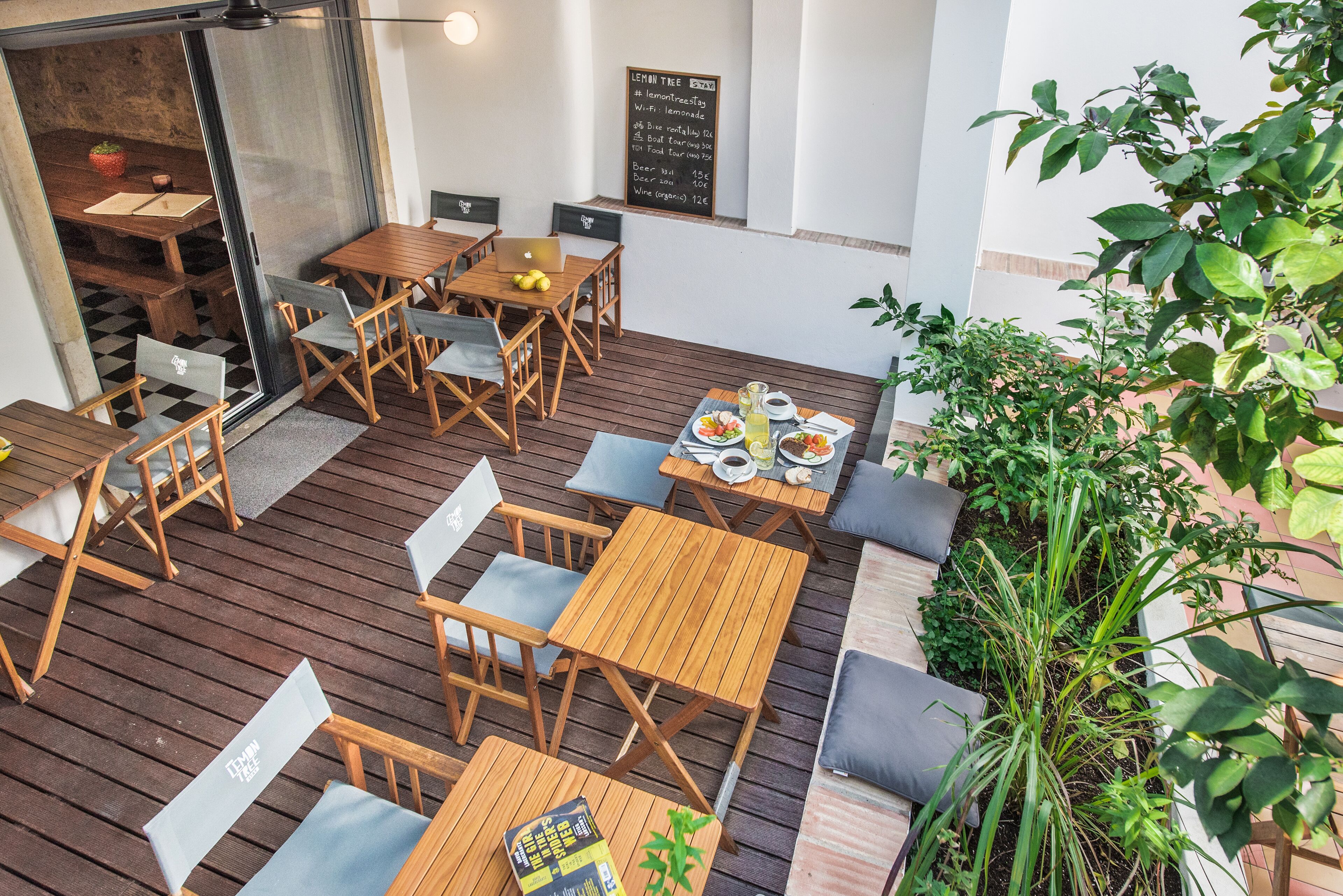 Un espacio al aire libre para comer con mesas y sillas de madera, un menú en una pizarra y plantas en macetas.