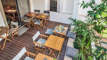 Un espacio al aire libre para comer con mesas y sillas de madera, un menú en una pizarra y plantas en macetas.
