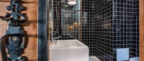 Ein Badezimmer mit weißem Waschbecken, schwarzen Fliesenwänden und einer Holztür mit Metallbeschlag.
