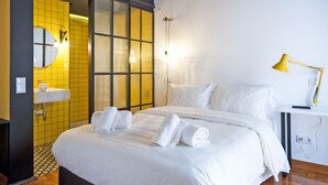 Una habitación de hotel con cama, una mesita de noche con lámpara, baño con lavamanos y espejo, y una puerta corredera que da a otra habitación.