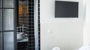 Una habitación de hotel con piso a cuadros, una cama con sábanas blancas, un televisor montado y un baño con baldosas blanco y negro.