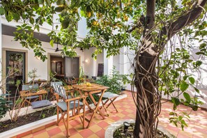 An outdoor seating area with wooden tables and chairs, surrounded by plants and a tree.