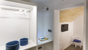 A room with a white wardrobe, two blue baskets, a white chair, a small table with a book, and a wall-mounted TV.