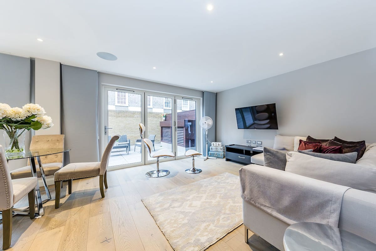 Ein modernes Wohnzimmer mit grauem Sofa, Couchtisch und Wand-Fernseher. Eine Glastür öffnet sich zu einem Außenbereich mit Stühlen und einem Tisch.
