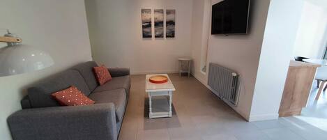 Una sala moderna con un sofá gris, una mesita blanca pequeña, un radiador y un televisor montado en la pared.