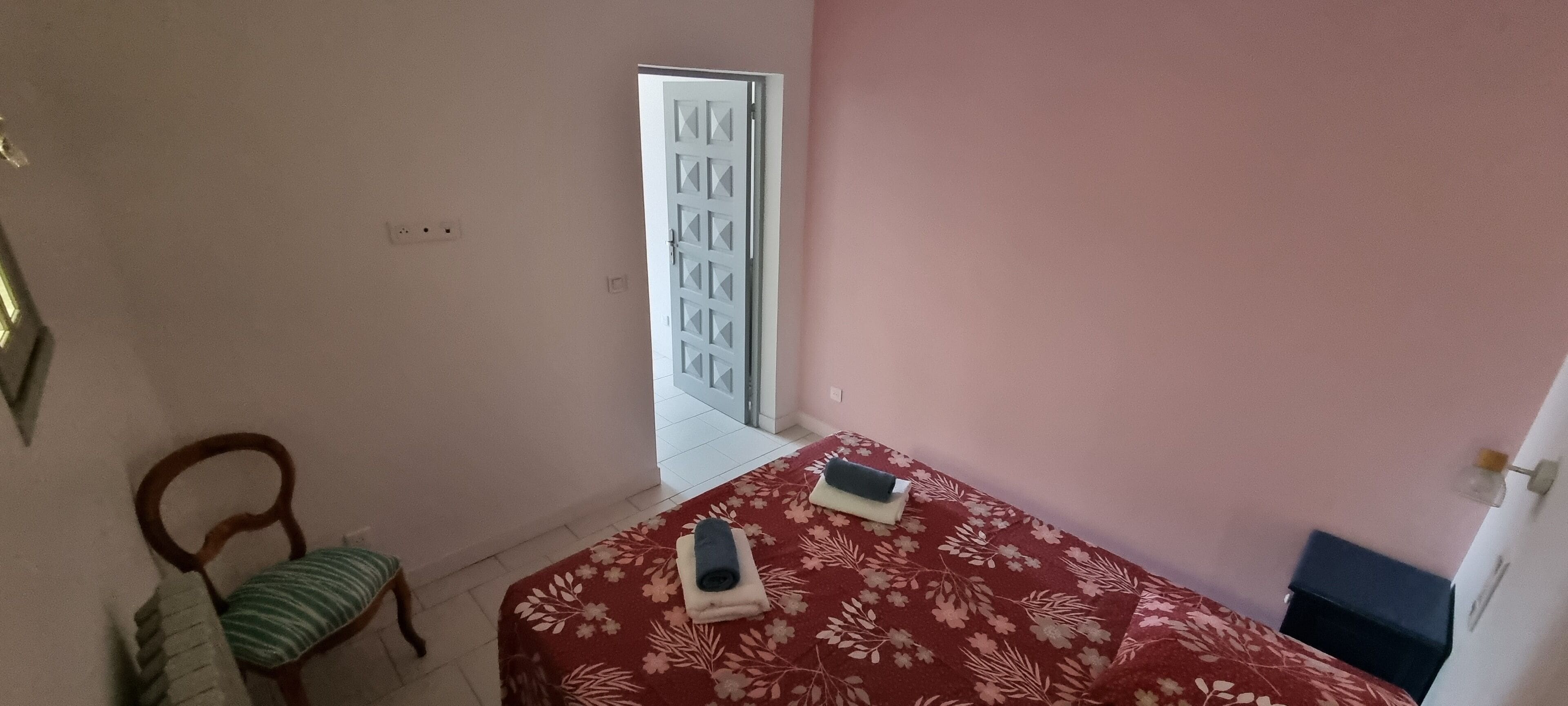 A bedroom with a bed covered in a floral-patterned bedspread, a chair with a striped cushion, and a door leading to another room.