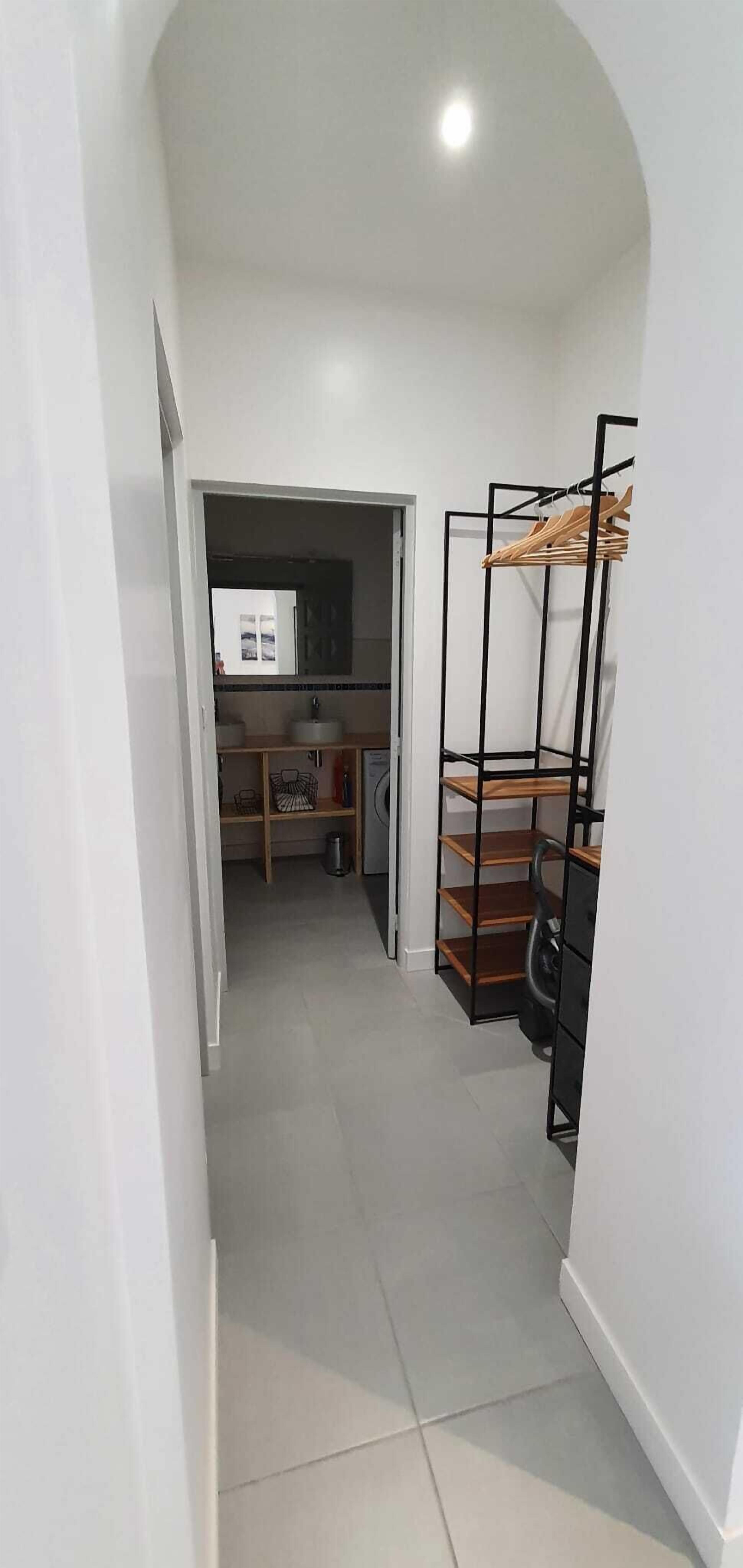 Un pasillo estrecho con una zona de kitchenette que incluye un lavamanos y armarios, una unidad de estanterías metálicas negras con repisas de madera y un piso de baldosas.
