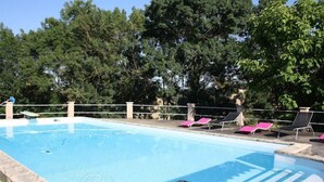 Una piscina rectangular con borde de piedra, rodeada por una cerca blanca. Hay sillones de descanso con cojines rosados a lo largo de la piscina.