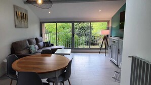 Una sala moderna con un sofá, una mesa de comedor de madera y sillas. Una ventana grande ofrece vistas al verdor exterior.