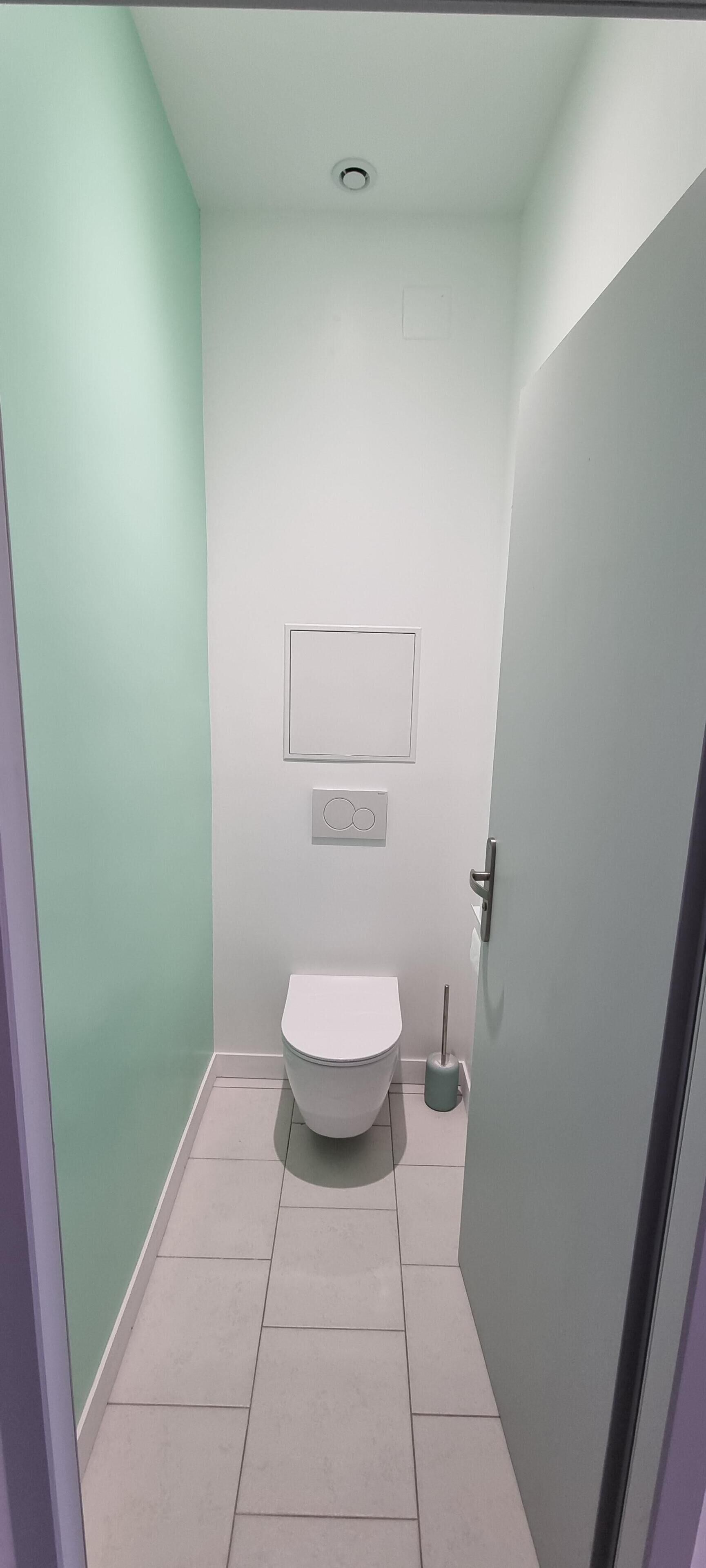 A narrow bathroom with a toilet, a hand dryer, and a wall-mounted light fixture.