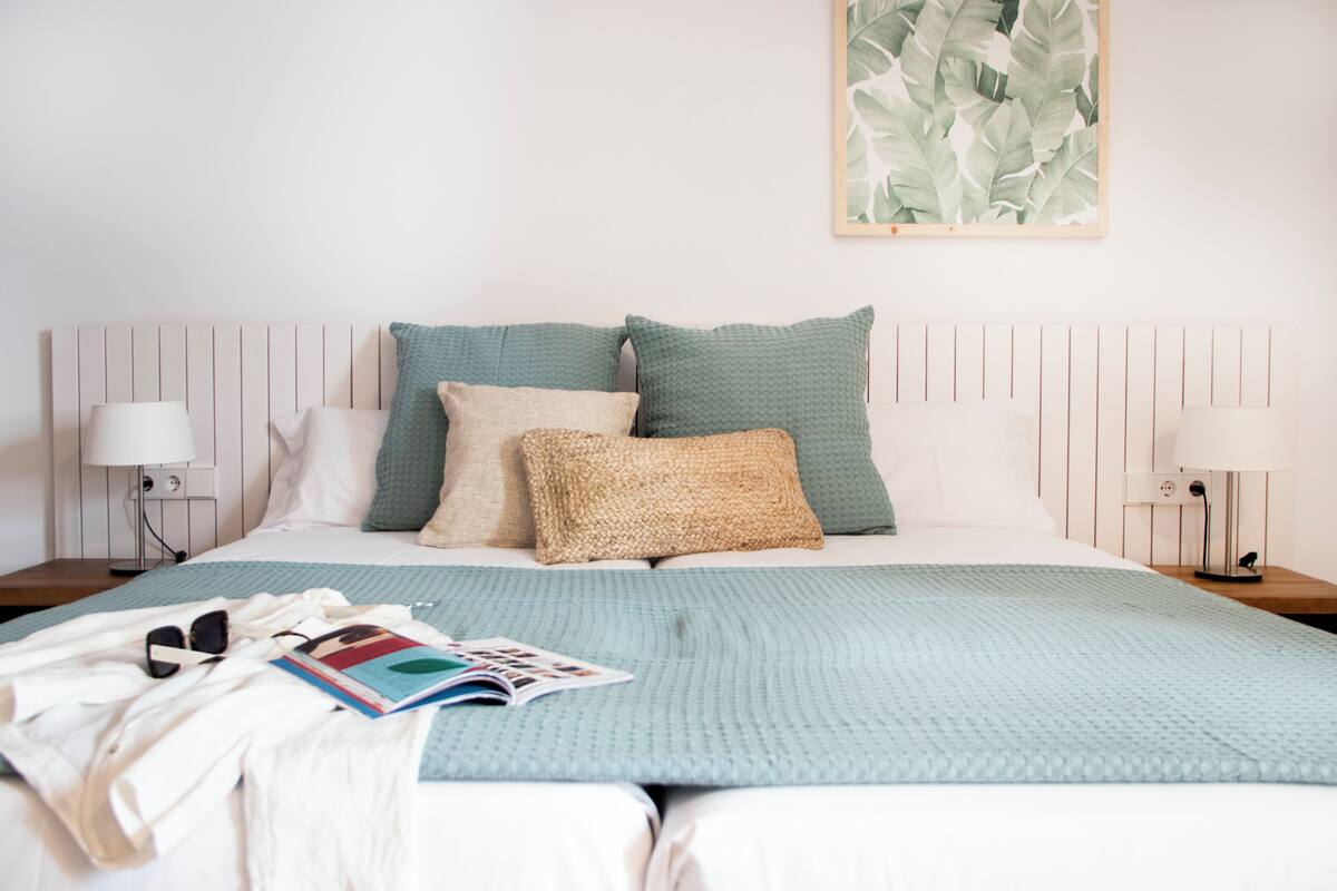 A neatly made bed with a light blue blanket, two pillows, and a magazine. There are two bedside tables with lamps and a framed plant above the bed.