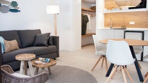 Een moderne woonkamer met een grijze bank, een ronde houten tafel, een rieten stoel en een witte eetkamerstoel. Achtergrond toont een kapstok en een houten plank met een lamp.