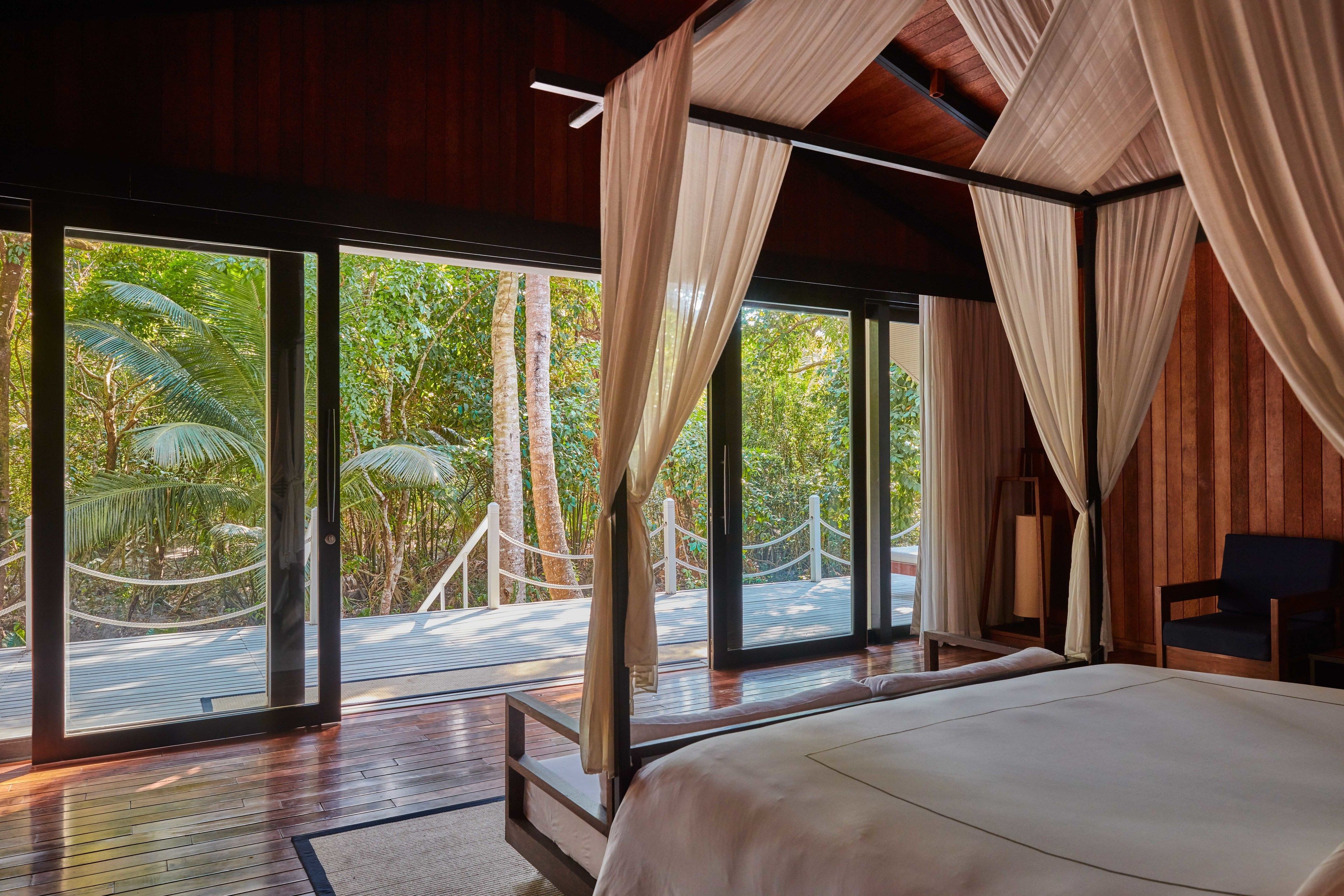 A bedroom with a large bed, wooden flooring, and a view of a lush garden through large windows.