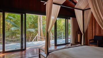 A bedroom with a large bed, wooden flooring, and a view of a lush garden through large windows.