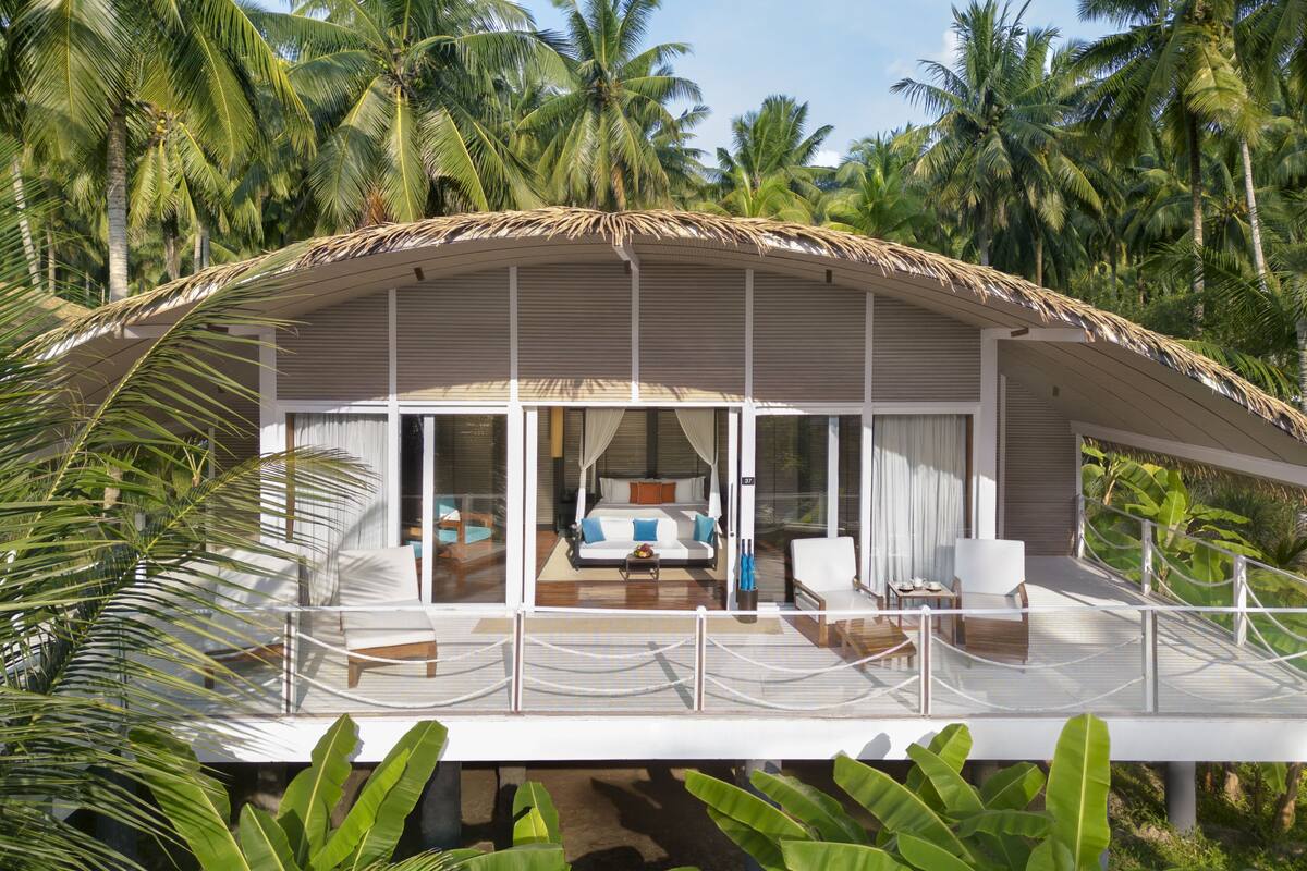 A modern, thatched-roof overwater bungalow with a spacious balcony, large windows, and a view of palm trees.