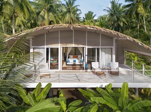 A modern, thatched-roof overwater bungalow with a spacious balcony, large windows, and a view of palm trees. - Taj Exotica Resort & Spa, Andamans (Port Blair)