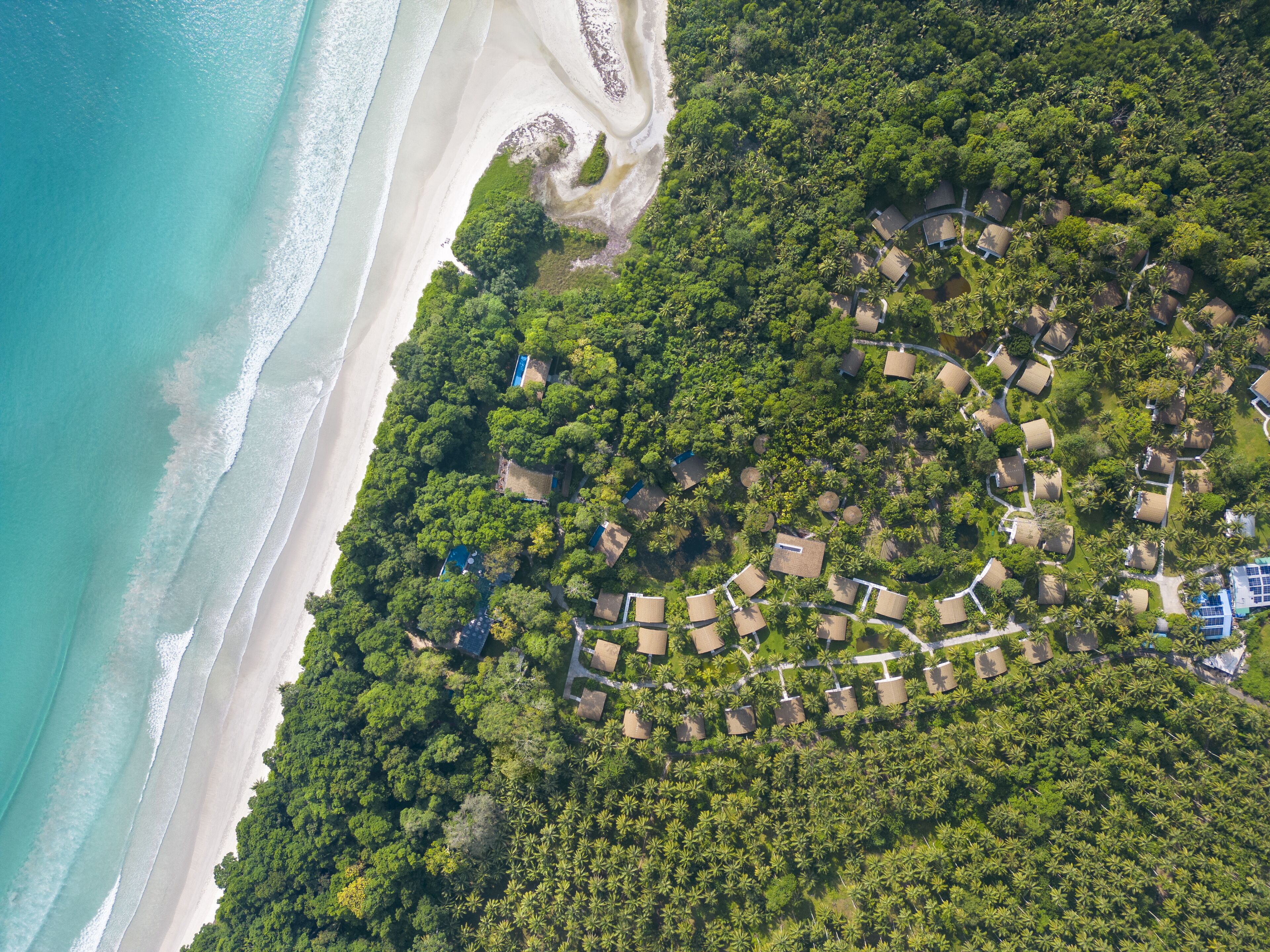 An aerial view of a resort with beachfront properties, dense greenery, and a clear view of the ocean.